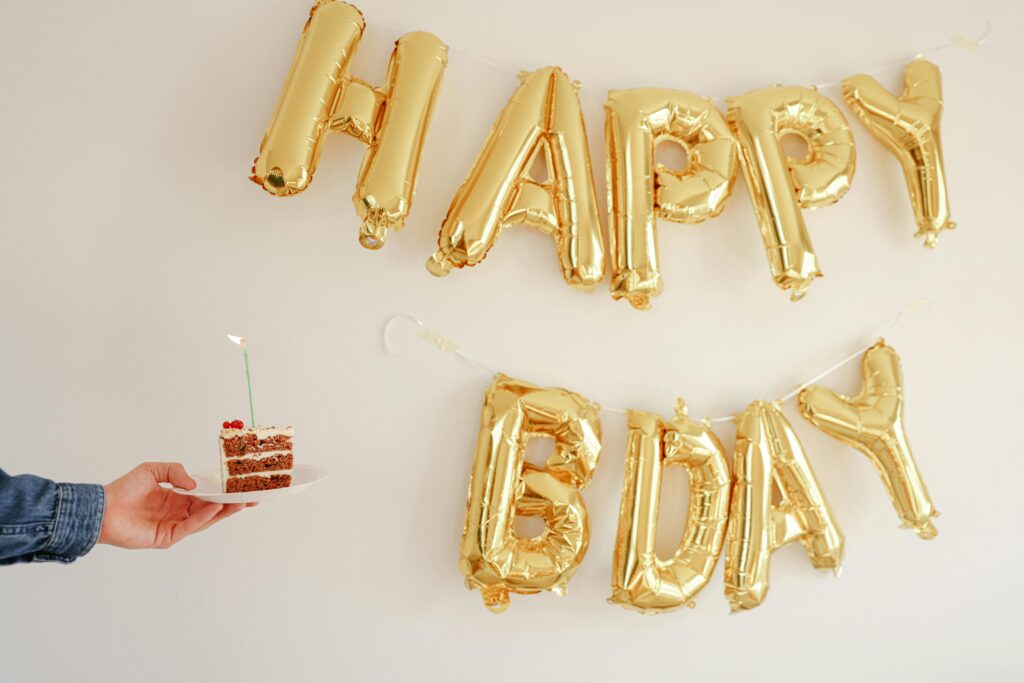 A slice of birthday cake with candle and gold 'Happy Bday' balloons on a white background.